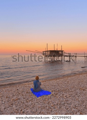 Punta Aderci, Vasto, Abruzzo, Italy: Young woman sitting on the beach of Adriatic sea at warm romantic sunset with a typical Mediterranean wooden structure fishing hut "Trabocco". Chieti