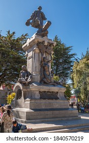 Punta Arenas, Chile - December 12, 2008: Ferdinand Magellan Statue. Bronze Sculptures On Gray Stone Pedestal Against Blue Sky With Green Foliage. People Hanging Around.