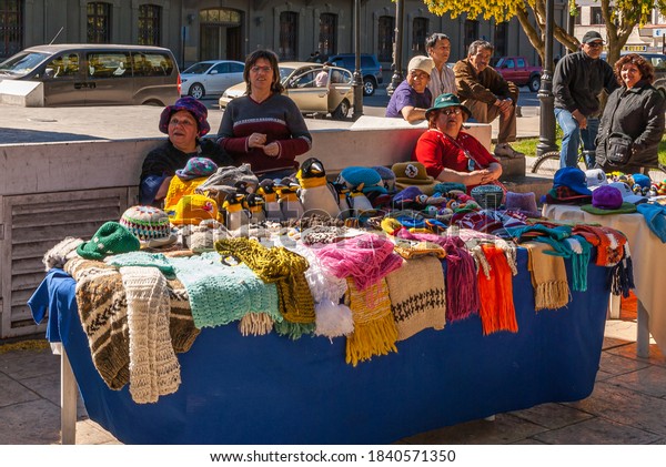 Punta Arenas, Chile - December 12, 2008: Ambulant\
female vendors sell colorful wool textiles off blue covered table\
along road with cars.