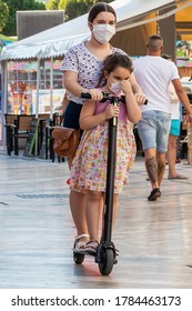 Punta Umbria, Huelva, Spain - July 10, 2020: Two Female Kids Wearing Protective Mask Due To Covid-19 Are Playing With A Electric Scooter. New Normal In Spain