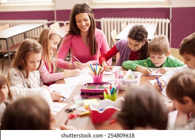 Pupil boys and girls sitting together around the table in classroom and drawing. With them is their young and beautiful teacher. She teaches children and smiling - Powered by Shutterstock