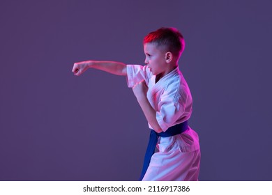 Pupil. Portrait Of Kid, Male Taekwondo, Karate Athletes In Doboks Doing Basic Movements Isolated On Purple Background In Neon. Concept Of Sport, Education, Skills, Martial Arts, Healthy Lifestyle.