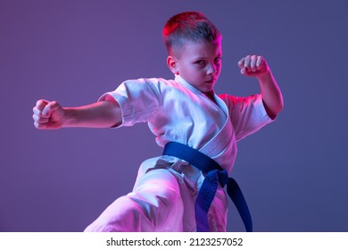 Pupil. Portrait of kid, male taekwondo, karate athletes in doboks doing basic movements isolated on purple background in neon. Concept of sport, education, skills, martial arts, healthy lifestyle.