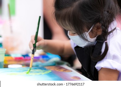 Pupils girl are concentrating on drawing and painting with brush and watercolor on the canvas. An Asian little child is wearing a white cloth face mask while learning to paint. Child 3 years old.