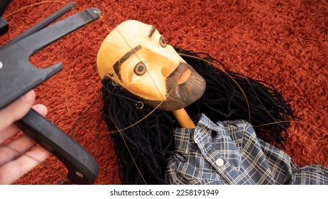 puppeteer hand with a crossbar tool of wood with a puppet or marionette hanging in the strings isolated over a red carpet background - traditional instrument for masters and a bald toy with beard - Shutterstock ID 2258191949