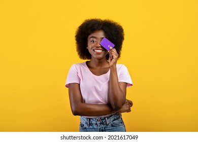 Purchase, money and payment concept. Joyful african american woman covering eye with credit card, wearing casual clothes posing isolated on yellow wall background, studio shot.