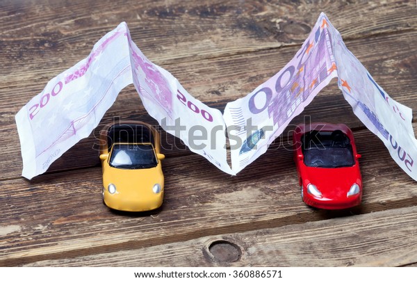 purchase, sale or car\
insurance.metaphor.Two little toy cars covered with paper money on\
a wooden surface