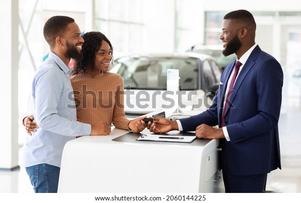 Purchasing Car. Happy Black Couple Taking Key\
From Manager In Dealership Center After Buying New Automobile,\
Young African American Spouses Choosing Auto In Modern Showroom,\
Smiling To\
Salesperson
