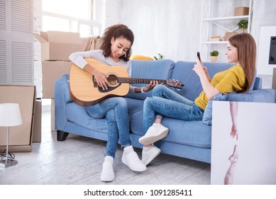 Pure Talent. Charming Young Girl Sitting On The Sofa Next To Her Roommate And Filming Her Playing Guitar