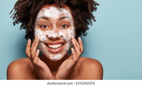 Purification of face. Happy smiling woman washes face with soap, cleans pores, touches cheeks, has spa procedures, uses gel, shows naked shoulders, isolated over blue background with blank space