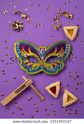 Purim celebration jewish carnival holiday concept. Tasty hamantaschen cookies, Triangular pastry, Festive carnival mask, noisemaker, sweet candies and party decor on purple Background, Top view.
