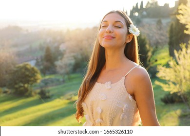 Purity And Beauty Concept. Portrait Of Beautiful Girl Breathing Fresh Air Outdoor In Nature With White Dress And Flower On Ear Smiling With Closed Eyes In Spring Summer Time At Sunset.