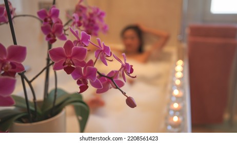 Purple blooming orchid with lined up glowing candles for creating relaxing atmosphere. Tranquil ambience for wellness treatment experience. Beautiful young woman enjoying and relaxing in bubbly bath. - Shutterstock ID 2208561423