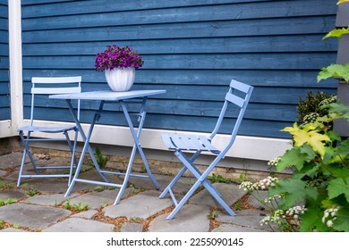 A purple colored metal patio table near a blue wooden horizontal clapboard siding exterior wall. There's a white flower pot with pink flowers on the small square table. The ground is patio flat rocks.