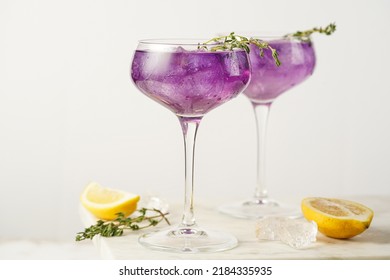 a purple drink in a vintage glass for sparkling wine - pea flower tea or blue curacao sirup cocktail with thyme branches on white background. Copy space for text