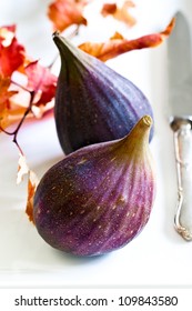 Purple figs on a white plate with a silver knife and colorful fall leaves.  Close up detail. Top view.