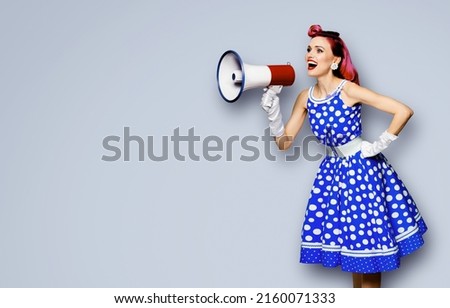 Purple haired woman holding red megaphone, shout advertising something. Girl in blue pin up style with mega phone loudspeaker. Grey color background with mock up. Female model in retro fashion dress.