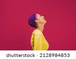 Purple haired young woman laughing cheerfully in a studio. Sideview of a fashionable young woman laughing with her eyes closed. Confident young woman standing against a pink background.