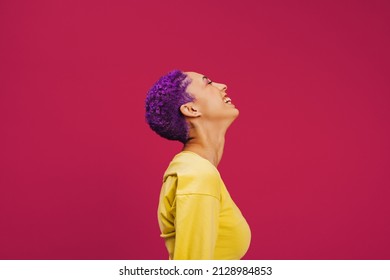 Purple haired young woman laughing cheerfully in a studio. Sideview of a fashionable young woman laughing with her eyes closed. Confident young woman standing against a pink background. - Shutterstock ID 2128984853