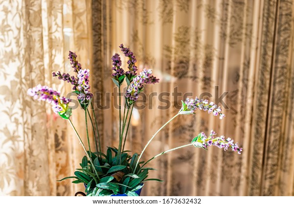 Purple lavender flowers bouquet arrangement inside\
vase potted plant closeup against window curtain blinds with fake\
cloth green leaves