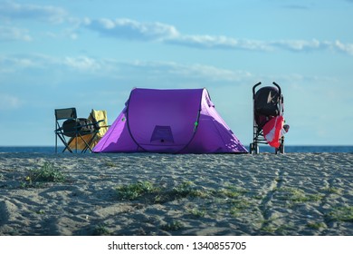 Purple Tent, Folding Camp Chair And Baby Stroller Are On The Sandy Beach