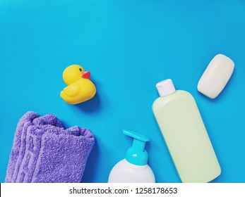 Purple Terry Towel, Yellow Rubber Duck, Liquid Soap, Shampoo And Soap. Flat Lay Baby Bath Products. Bathroom Items On A Blue Background