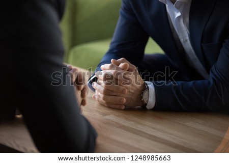 Purposeful confident restrained businessmen in suits sit at table opposite each other, close up male clenched hands in lock at desk. Complexity of decision self-control in stressful situation concept