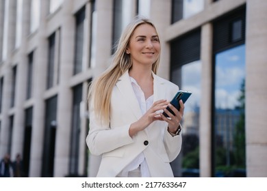 Purposeful young caucasian blonde woman in white suit standing outdoor with phone on summer shiny day. Happy student swedish girl going to call her boyfriend. Cute smiling Italian pretty woman.