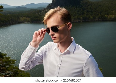 A purposeful young man in a white shirt and black sunglasses stands against a lake and mountain landscape. Business concept. Vacation and travel.