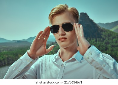 A Purposeful Young Man In A White Shirt And Black Sunglasses Stands Against A Mountain Landscape. Business Concept. Vacation And Travel.