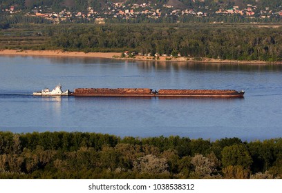 The Pusher Vessel Tows A Train Of Two Barges Laden With Logs, Along A Large River In Clear Weather