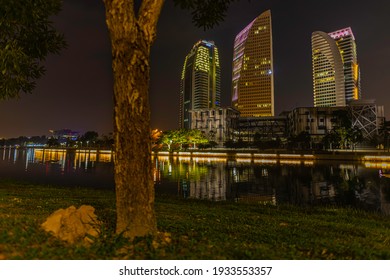 Putrajaya, Malaysia - 2021, February 26: Time Lapse Close To The Ground With The Green Grass. View To Office Buildings Near The Putra Lake. Tree Out Of Focus In The Foreground. Sunset Behind Buildings