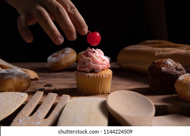 Putting a cherry on a top of pink cup cake among its ingredients with dark background, wooden spoon and fork. - Powered by Shutterstock