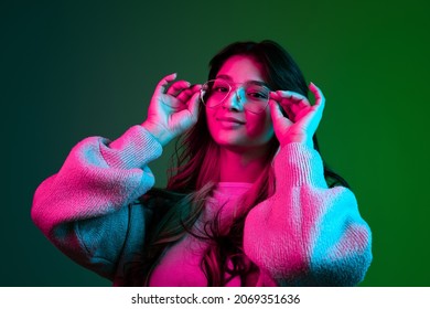 Putting eyeglasses on. Beuatiful Latino cute girl, student in casual style clothes posing isolated on dark green studio backgroud in pink neon light. Emotions, facial expression, youth, fashion and ad