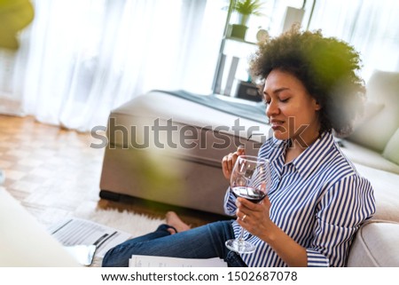 Putting fresh content into her blog. Thoughtful young woman in eye-wear working using computer while flooring at home with glass of wine. Wine brings fresh ideas. 