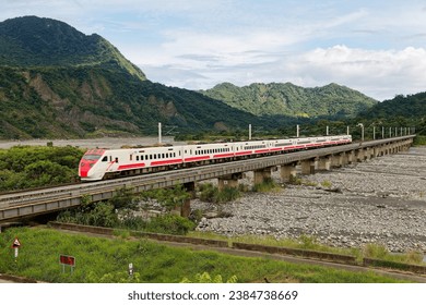 A Puyuma express train dashing thru a bridge over the stony, dry riverbed of Luye River 鹿野溪, with majestic mountains under blue sunny sky in the background, in Beinan Township, Taitung County, Taiwan