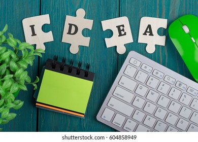 Puzzle pieces with text "idea", blank note pad, computer keyboard and mouse on a blue wooden background