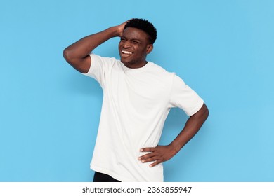 puzzled african american guy in white t-shirt remembers and scratches his head on blue isolated background, confused man forgot and thinks