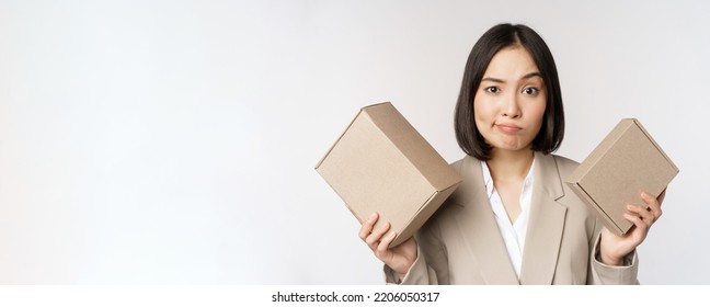 Puzzled Asian Saleswoman Holding Boxes, Looking Confused, Standing In Business Suit Over White Background