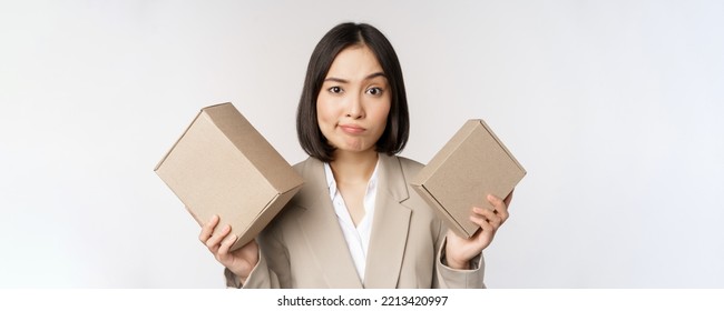 Puzzled Asian Saleswoman Holding Boxes, Looking Confused, Standing In Business Suit Over White Background