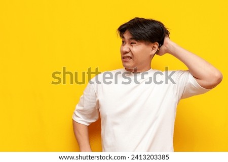puzzled asian young man in white t-shirt thinks and remembers over yellow isolated background, confused korean guy student forgot and scratches head looking away