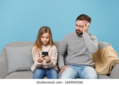 Puzzled Bearded Man In Knitted Sweater With Child Baby Girl. Father Little Daughter Isolated On Pastel Blue Background. Love Family Parenthood Childhood Concept. Sitting On Couch, Hold Mobile Phone