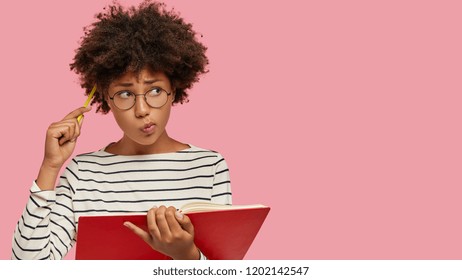 Puzzled Black Woman Has Contemplative Expression, Writes To Do List Of Goals, Holds Notebook, Scratches Head With Pencil, Looks Aside, Being Deep In Thoughts, Isolated On Pink Studio Background