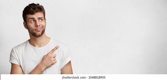 Puzzled blue eyed handsome young bearded man poses agianst blank copy space, dressed casaully, indicates with fore finger aside, raises eyebrows in bewilderment. Advertisment and people concept