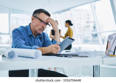 Puzzled Businessman In Optical Eyewear Reading Financial Publication Browsed On Modern Touch Pad Using Office Internet Connection At Table Desktop, Caucasian Executive Manager Checking Email Text