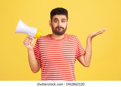 Puzzled Confused Man With Beard In Striped T-shirt Shrugging Shoulders Holding In Hands Megaphone, Cant Find Common Language, Communication Problems. Indoor Studio Shot Isolated On Yellow Background