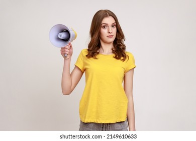 Puzzled Confused Teenager Girl With Brown Hair In Yellow T-shirt Holding In Hands Megaphone, Cant Find Common Language, Looking At Camera. Indoor Studio Shot Isolated On Gray Background.