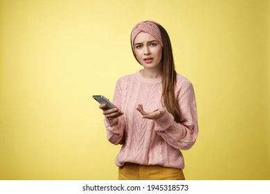 Puzzled confused upset questioned young cute girl in headband knitted sweater frowning clueless raising hand in dismay holding smartphone, cannot understand what happened cellphone over yellow wall - Shutterstock ID 1945318573