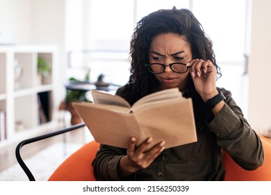 Puzzled confused young black woman reading book and taking off or putting on eyeglasses sitting on pouf bean bag chair, penisve lady spending time alone in living room at home. Incomprehension