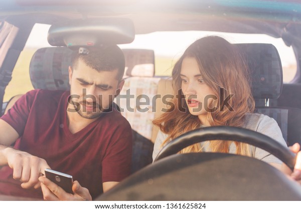 Puzzled couple sits in automobile, bearded man\
holds smart phone, uses online maps, tries to find way, being lost,\
have journey on weekend. Family in automobile chek email while\
parking on road side.